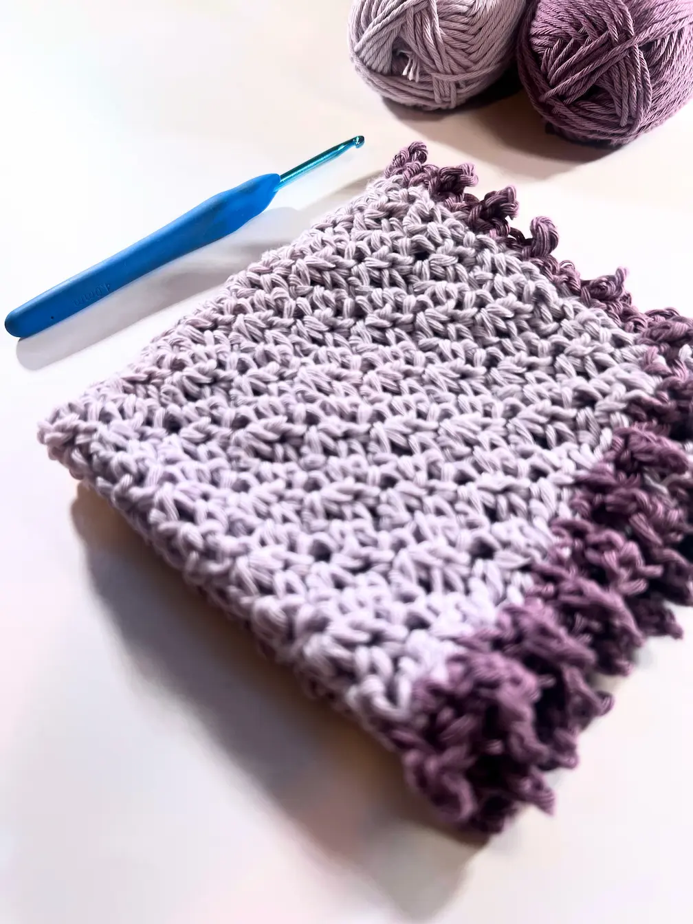 Folded crochet washcloth with a purple picot border, placed next to a blue crochet hook and two yarn balls in lavender and dark purple.