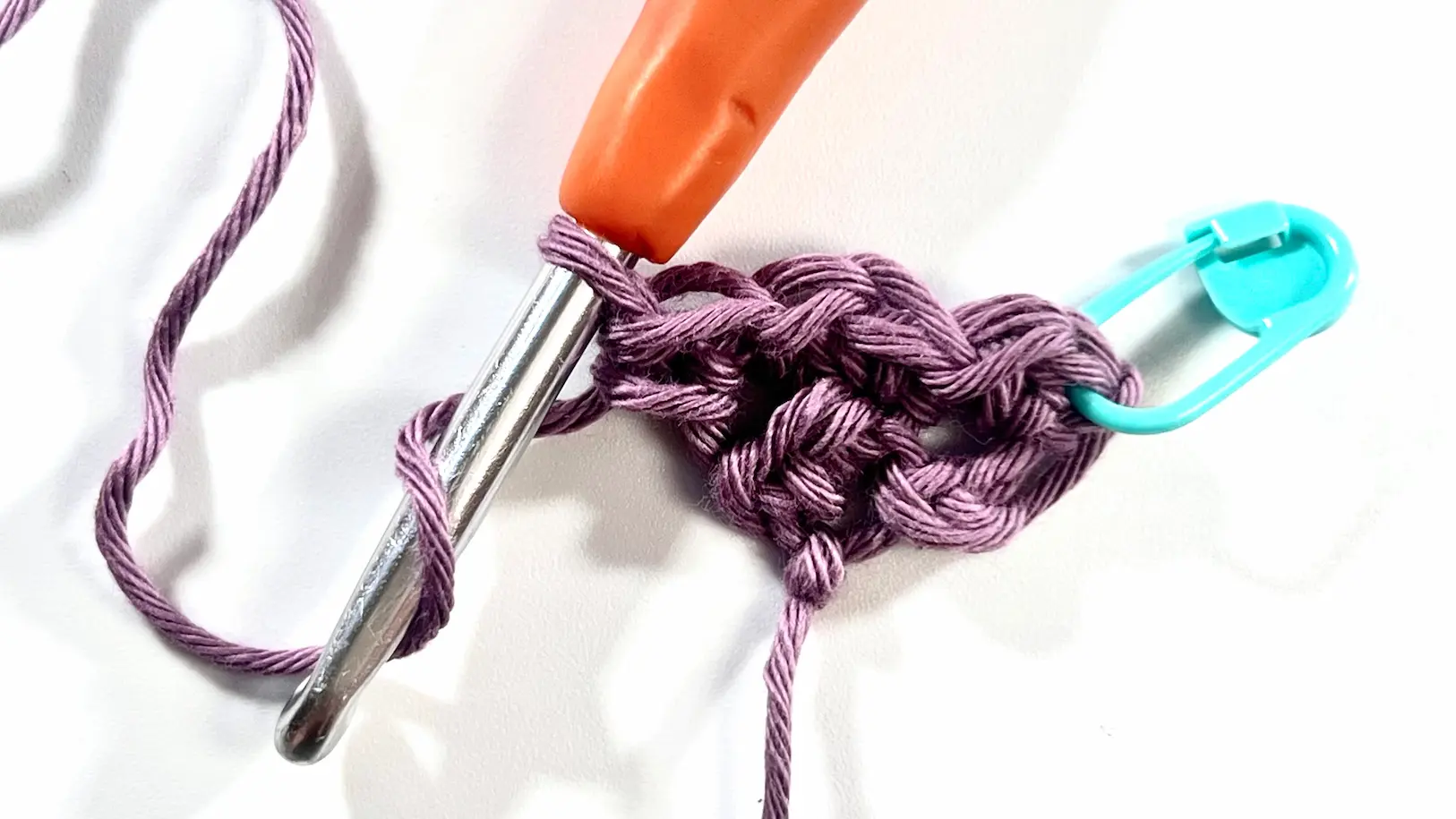 Two full corner-to-corner blocks completed in Even Moss Stitch using purple cotton yarn. The crochet hook is ready to continue with the next row, and a teal stitch marker highlights the base of the row.