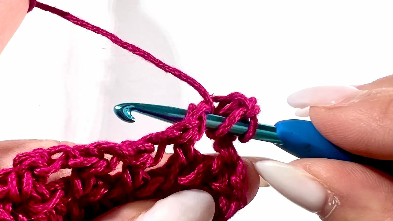 Close-up showing how to crochet the feather stitch step-by-step using fuchsia yarn and a blue crochet hook — part of a textured stitch tutorial.