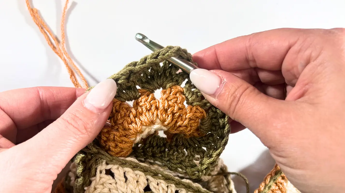 Close-up of a crochet hook inserted into the corner chain space of a green daisy granny square, ready to start the strap.