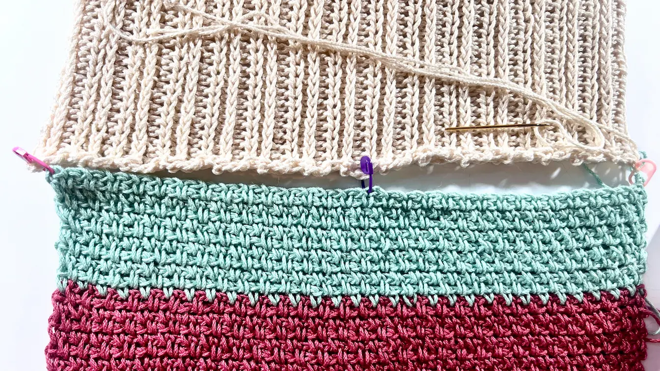 Two crochet sweater panels laid flat and aligned with the help of colourful stitch markers, showing a yarn needle threaded and ready for seaming at the shoulder edge.