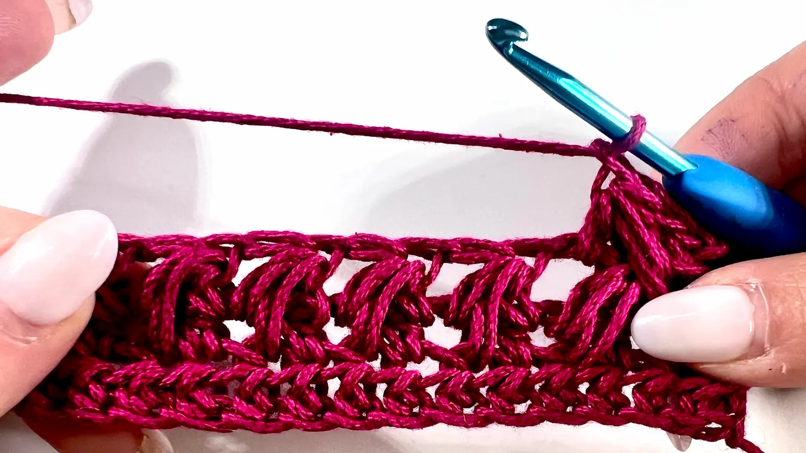Close-up showing how to crochet the feather stitch step-by-step using fuchsia yarn and a blue crochet hook — part of a textured stitch tutorial.