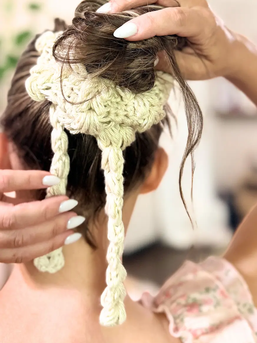 Cream crochet Blooming Heart Scrunchie with puff stitch petals and braided ties styled around a messy bun hairstyle.