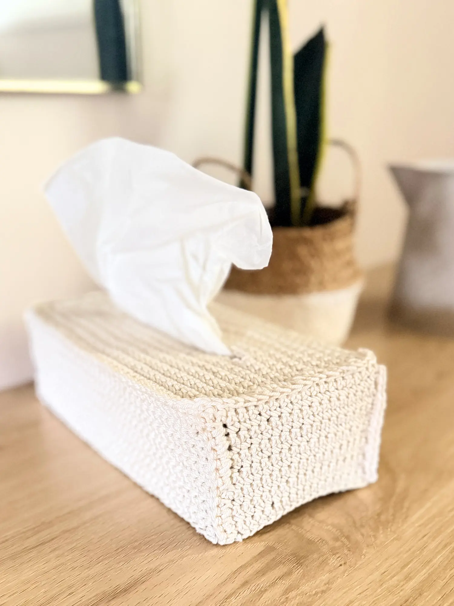 Modern crochet tissue box cover displayed on a wooden table with a tissue box inside and a tissue pulled through the opening.