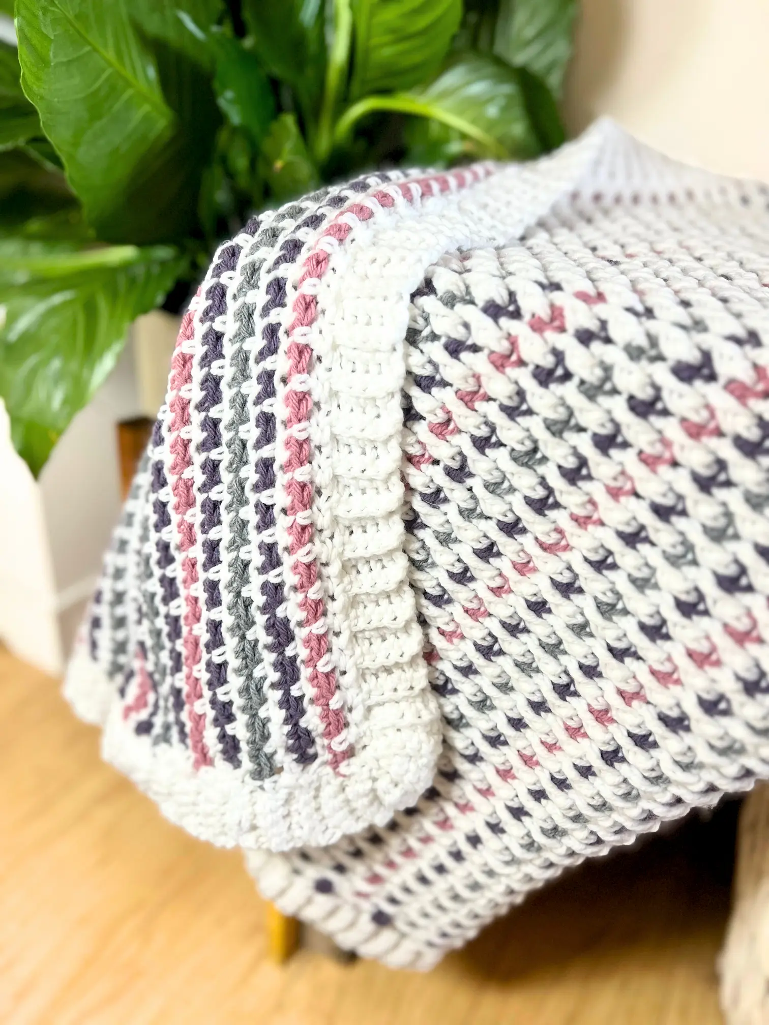 Cuddle Blanket draped over a chair, showing textured Alpine Stitch rows in white, pink, grey, and purple with a ribbed border.