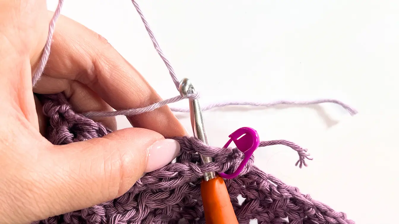The first seam stitch has been completed to join the hood and towel together. The crochet hook is pulling up a loop of dusty lavender yarn near a pink stitch marker securing the layers.