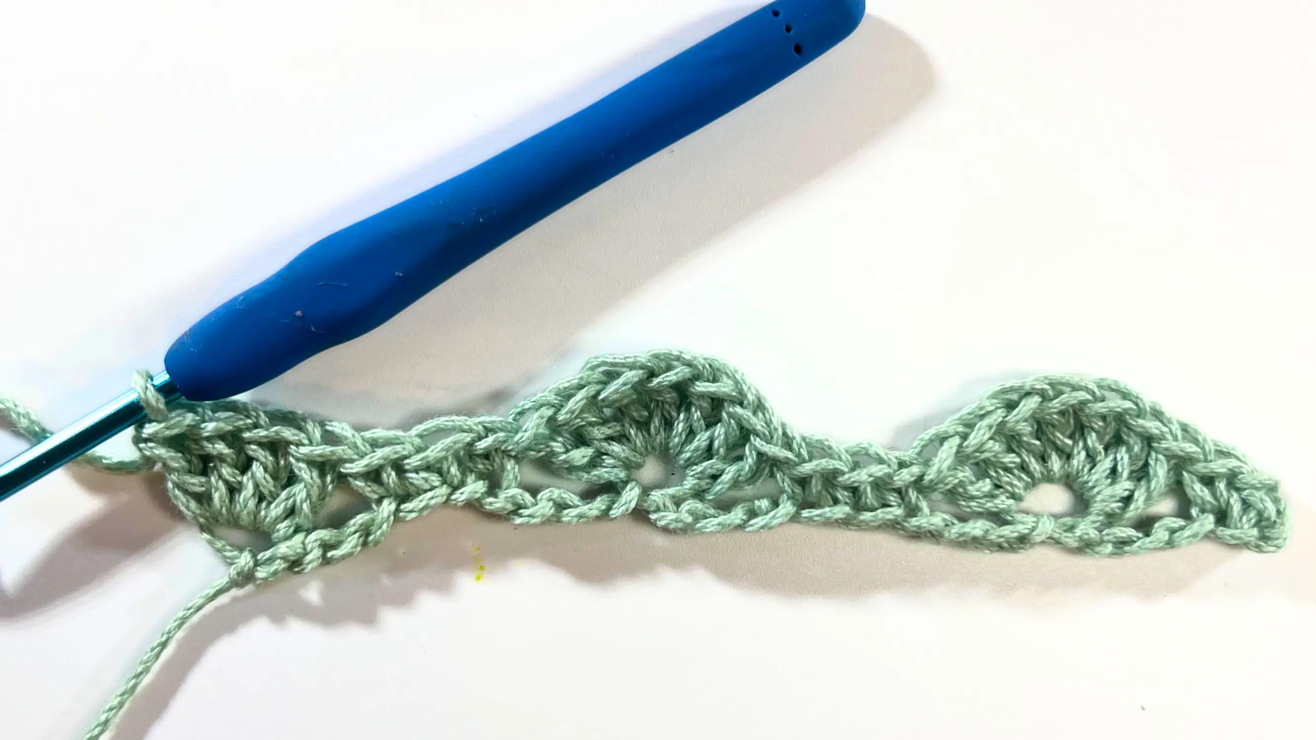 Row 1 of the Catherine Wheel Stitch worked in light green yarn, showing two completed 7 double crochet shells and a 4 double crochet shell at the end. A blue crochet hook is attached on the left side of the row.
