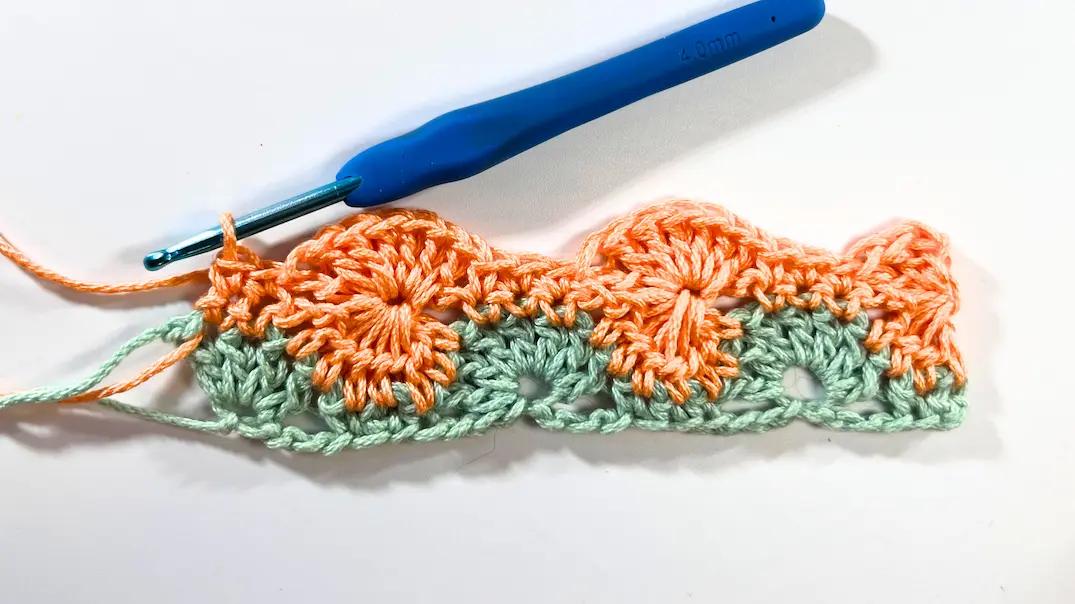 Completed three rows of the Catherine Wheel Stitch worked in green and orange yarn. The wheel motifs are fully visible with scalloped edges across the row.