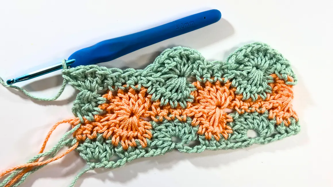 Sample of the Catherine Wheel Stitch with five rows completed in alternating green and orange yarn, showing the full wheel pattern design with a crochet hook resting at the top.