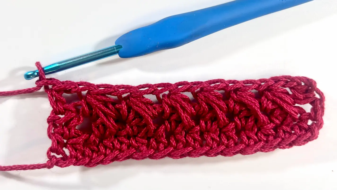 Close-up showing how to crochet the feather stitch step-by-step using fuchsia yarn and a blue crochet hook — part of a textured stitch tutorial.
