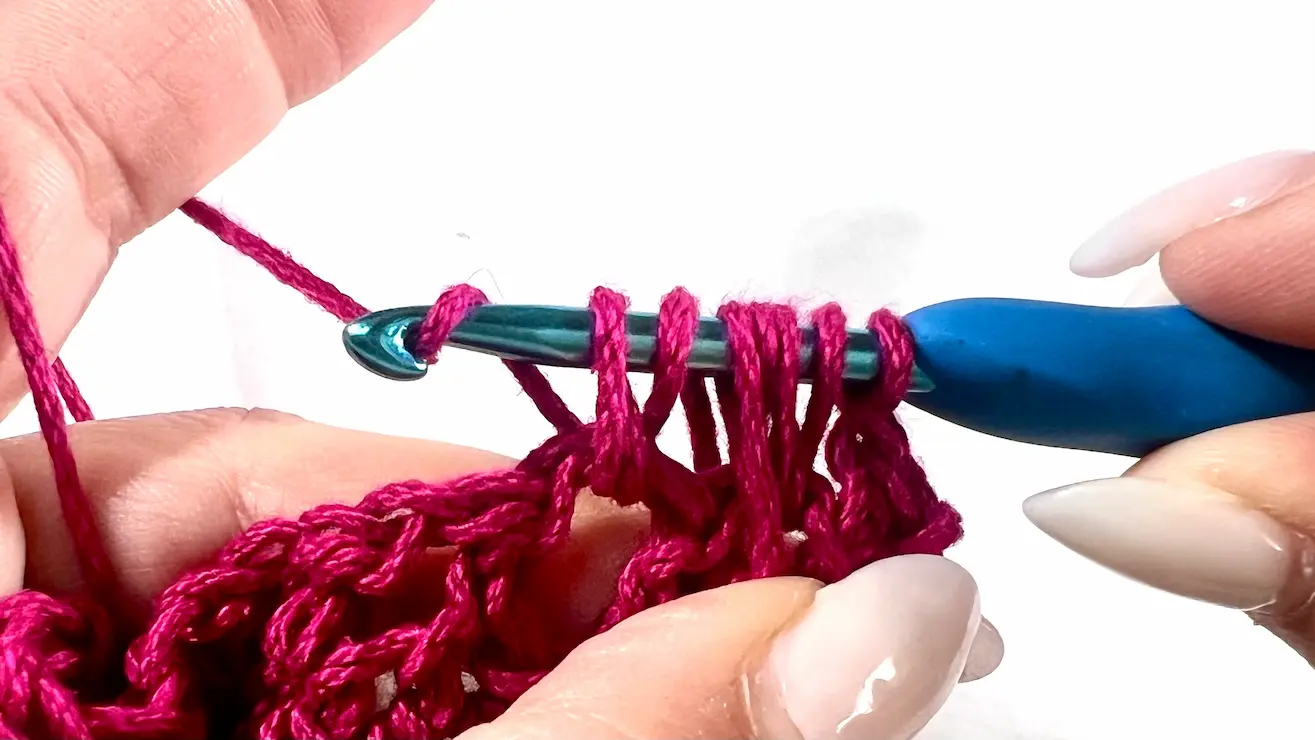 Close-up showing how to crochet the feather stitch step-by-step using fuchsia yarn and a blue crochet hook — part of a textured stitch tutorial.