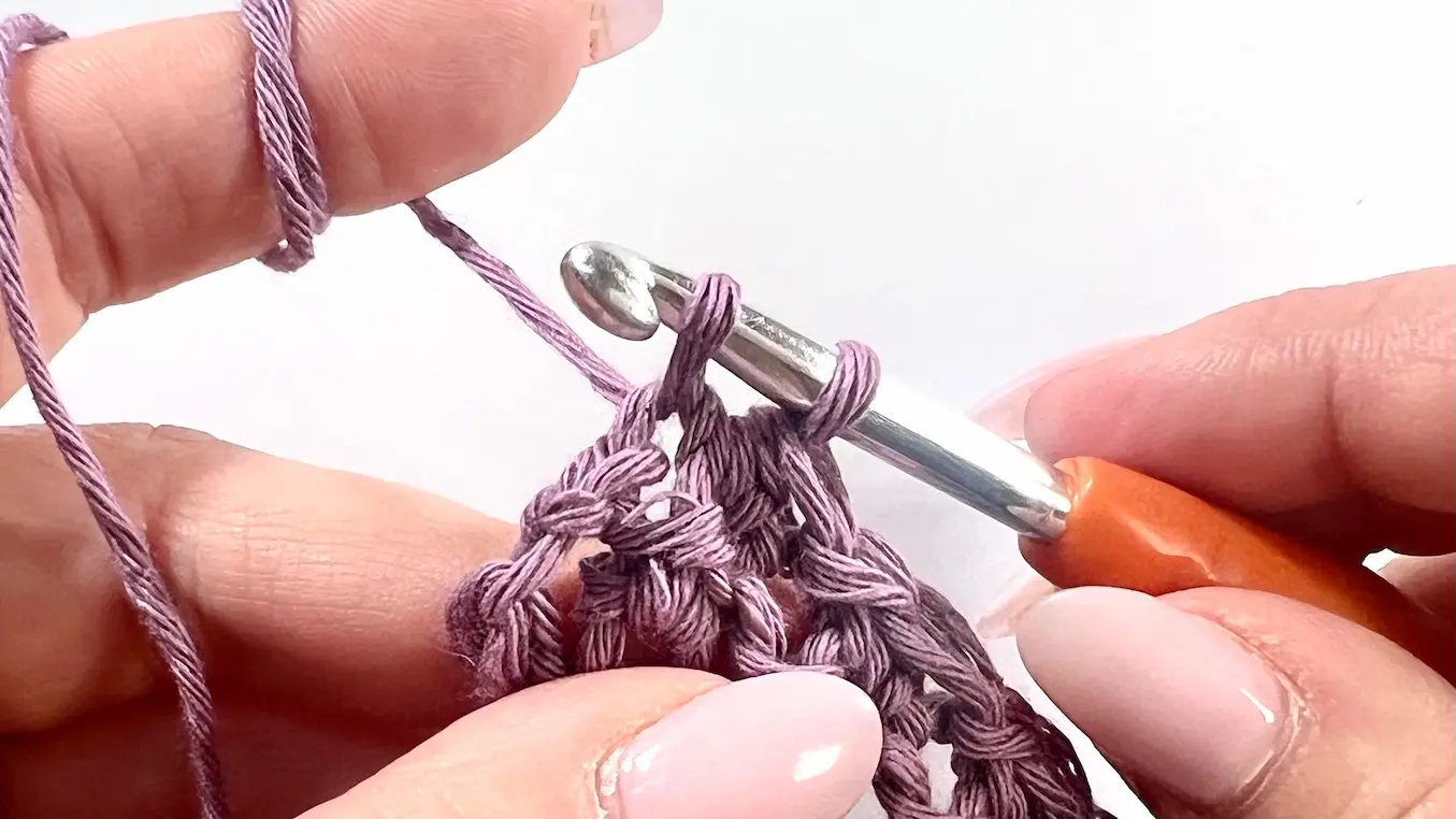 Crochet hook with two loops, about to complete a half double crochet. The yarn is light purple cotton and the stitch is being worked into a marked space as part of a C2C decrease.