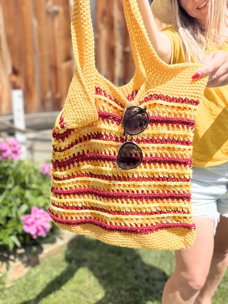 Handmade crochet summer beach tote bag with sturdy handles, textured stripes, and sunglasses hanging from the front, held outdoors in the sunshine.