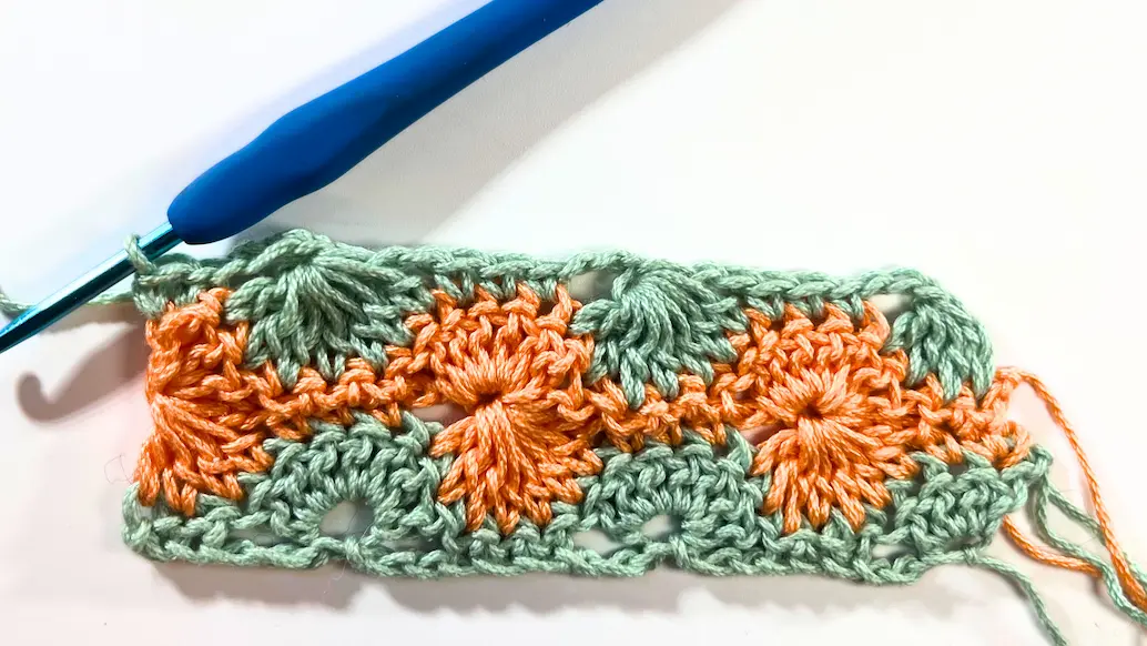 Progress shot of the Catherine Wheel Stitch showing several repeats of 7DC together clusters and 3 SC groups, worked in alternating rows of orange and green yarn.