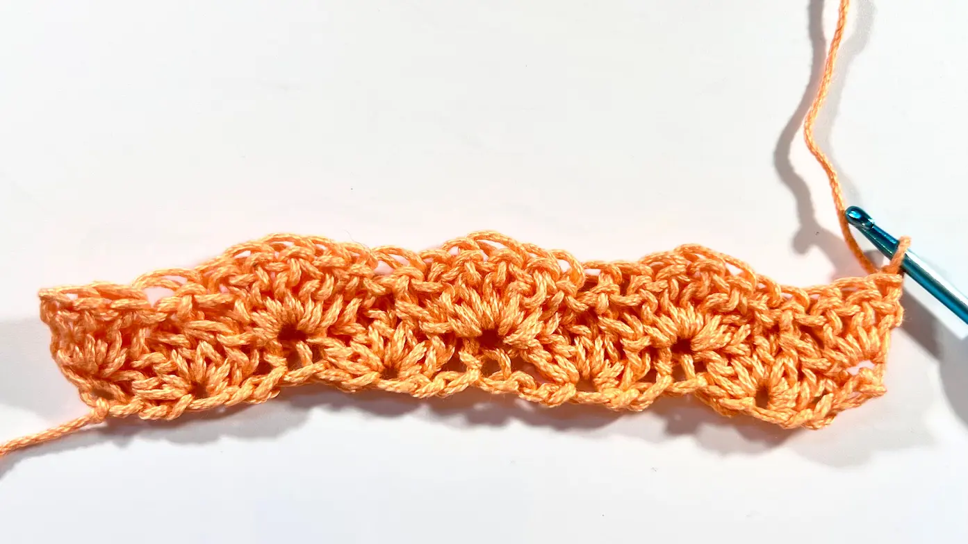 A completed swatch showing two full rows of shell stitch in orange yarn, with a blue crochet hook attached and ready to continue.