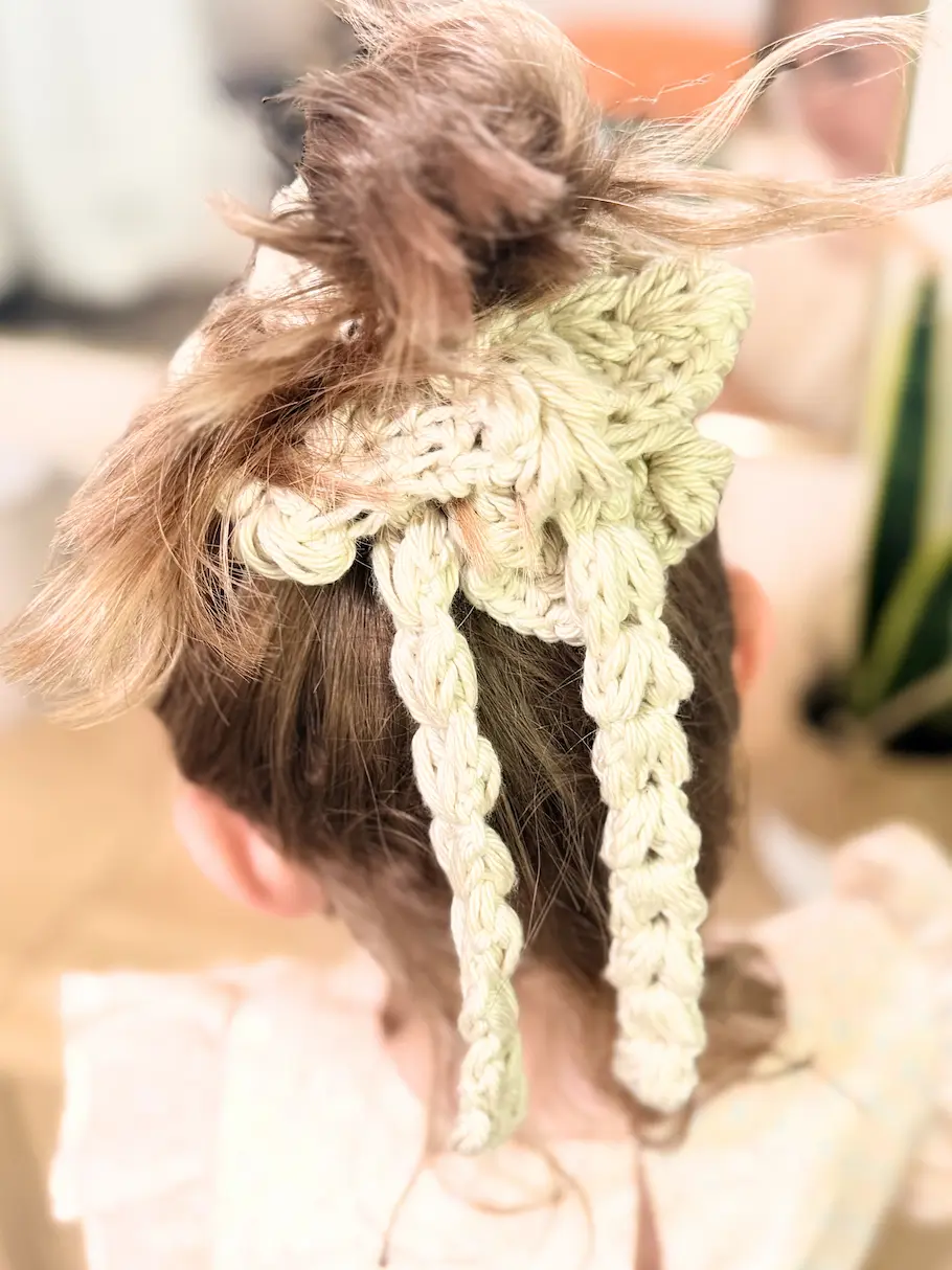 Back view of a cream crochet scrunchie featuring textured puff stitches and two long braided ties wrapped around a bun.