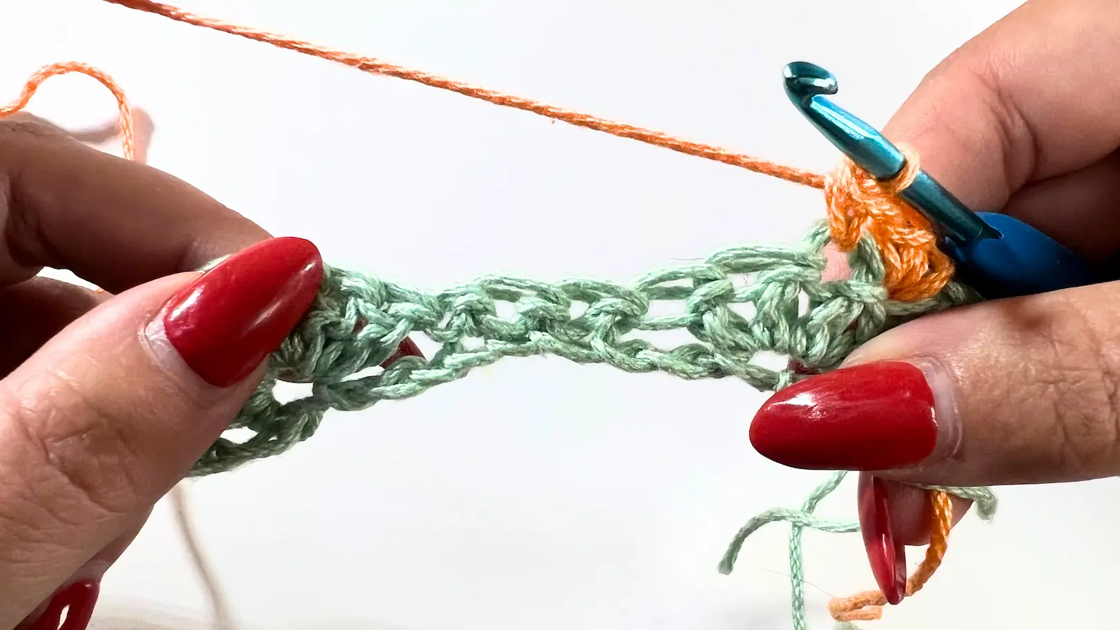Orange yarn worked into the green row to begin Row 2 of the Catherine Wheel Stitch. The blue crochet hook is inserted into the stitch, with red nails holding the work in place.