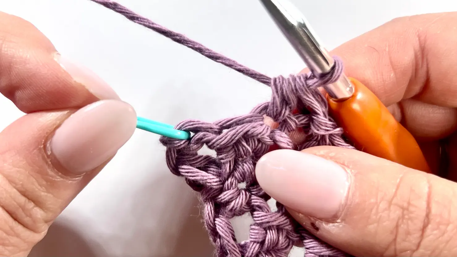 Close-up of a C2C Even Moss Stitch crochet project in progress. A teal stitch marker is inserted into the last stitch of the block to indicate where the final stitches of the decrease should go. The orange-handled hook is positioned above the stitch.