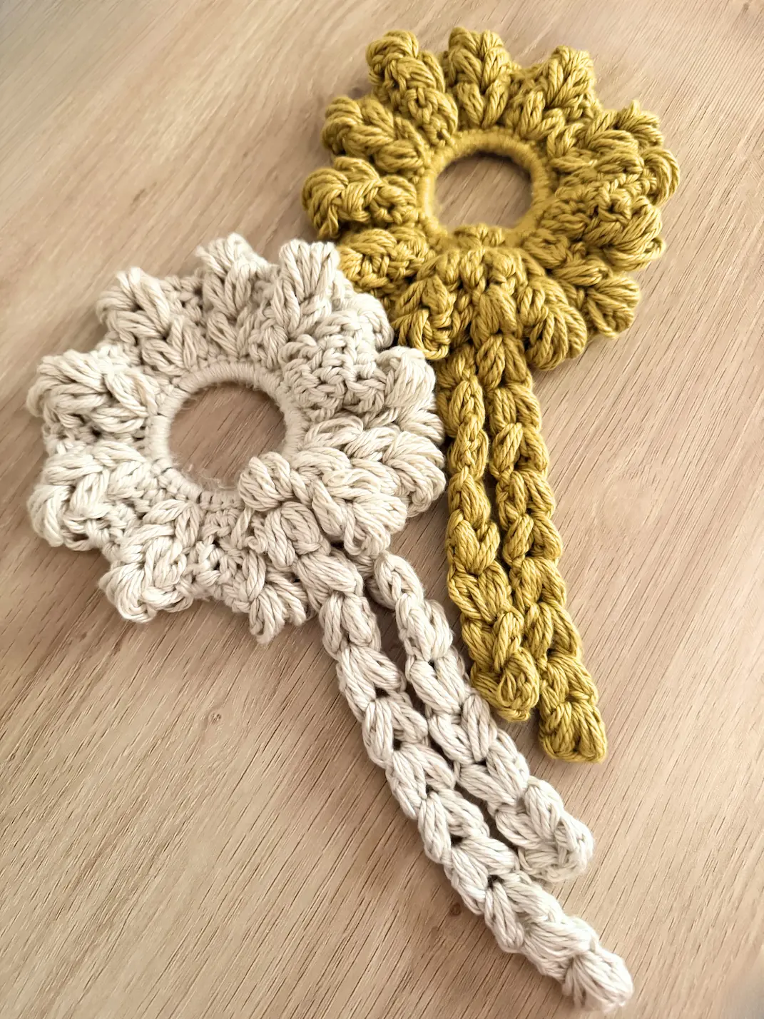 Cream and mustard crochet Blooming Heart Scrunchies with puff stitch petals and long braided ties displayed on a wooden surface.