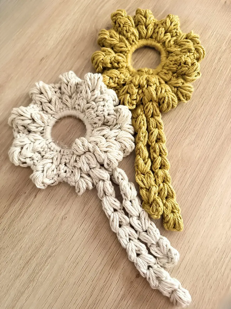 Cream and mustard crochet Blooming Heart Scrunchies with puff stitch petals and long braided ties displayed on a wooden surface.