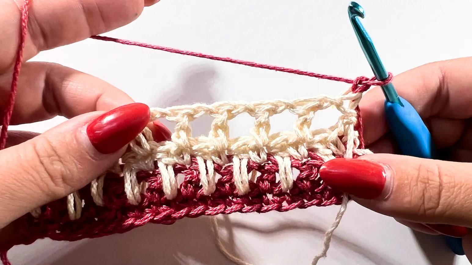 Crochet swatch with four completed rows, alternating cream and red, showing the spike stitch texture with hook positioned at the top ready for the next row.