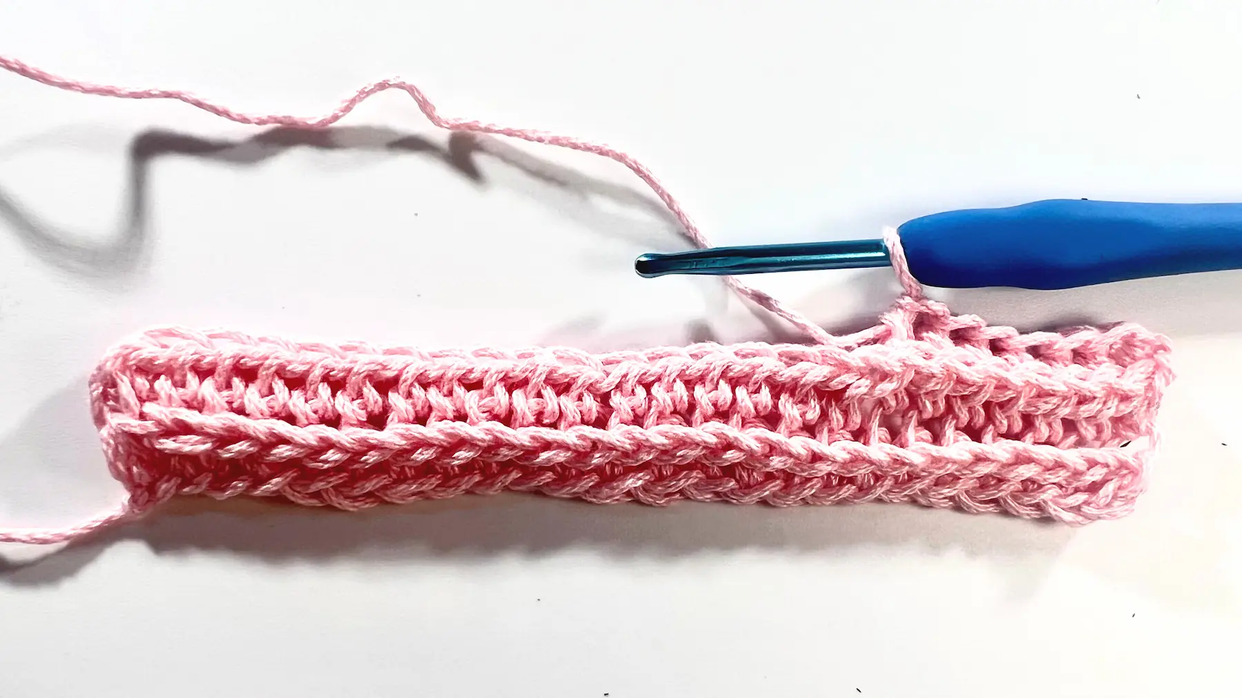 Progress view of a crochet swatch in pink yarn with three rows of camel stitch completed, showing distinct ridges forming across the fabric.