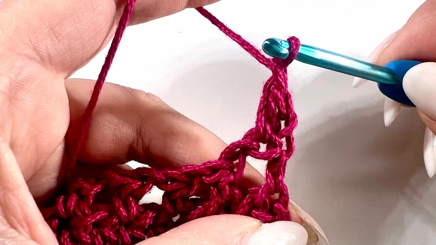 Close-up showing how to crochet the feather stitch step-by-step using fuchsia yarn and a blue crochet hook — part of a textured stitch tutorial.