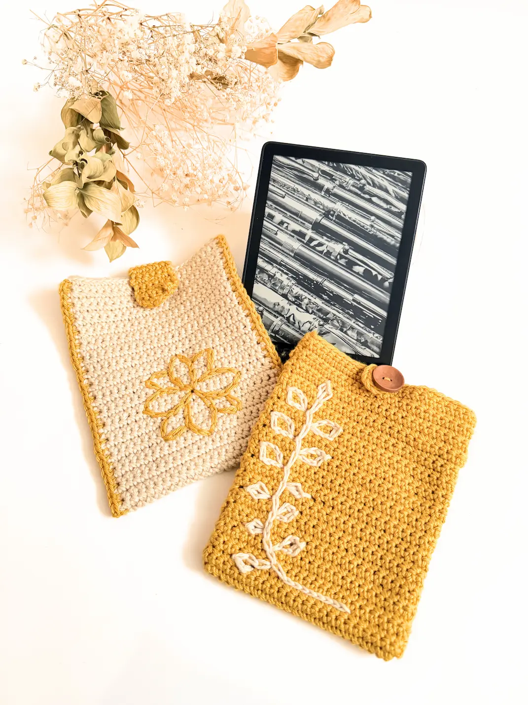 Two crochet Kindle sleeves displayed beside an e-reader, featuring a floral surface crochet design and a leaf surface crochet design in cream and mustard yarn.