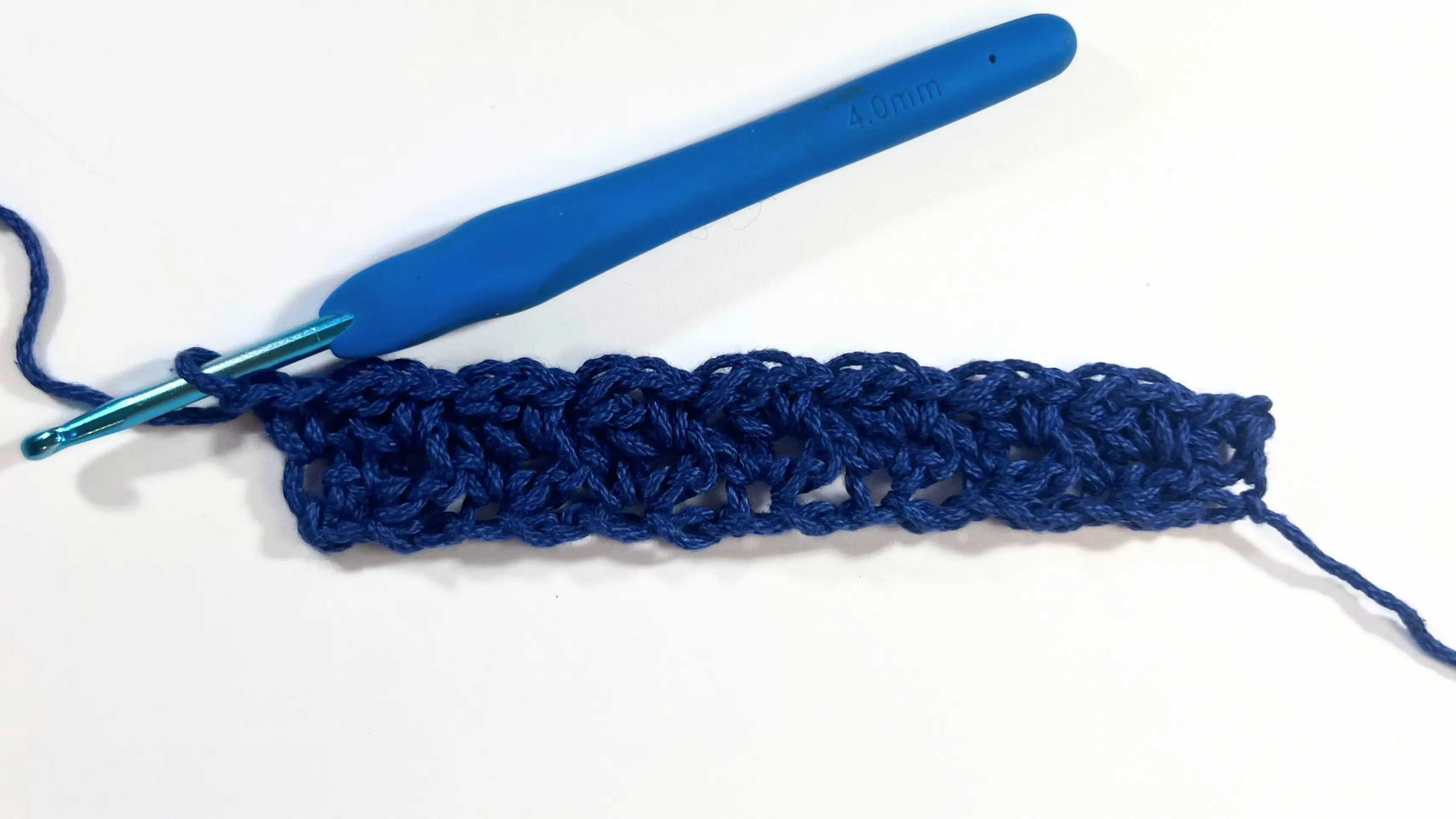 Two full rows of wattle stitch completed in blue yarn with a 4.0mm blue crochet hook attached on the left side, ready for the next row.