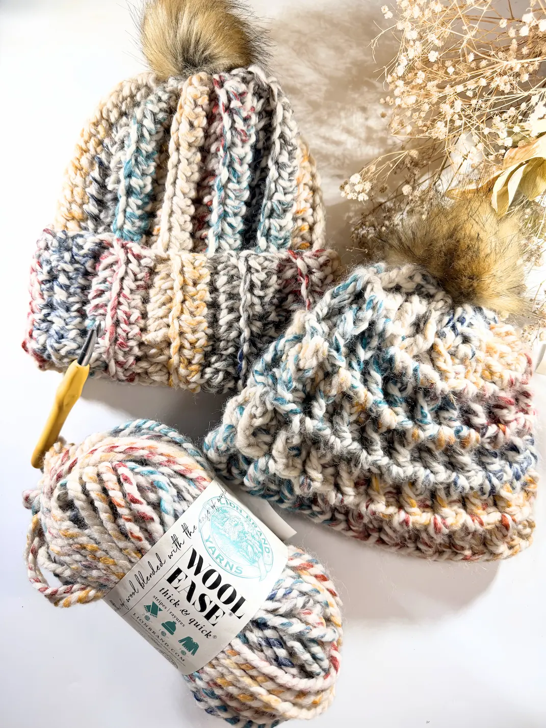 Two finished chunky crochet beanies with faux fur pompoms displayed beside Lion Brand Wool-Ease Thick & Quick yarn and crochet hook.