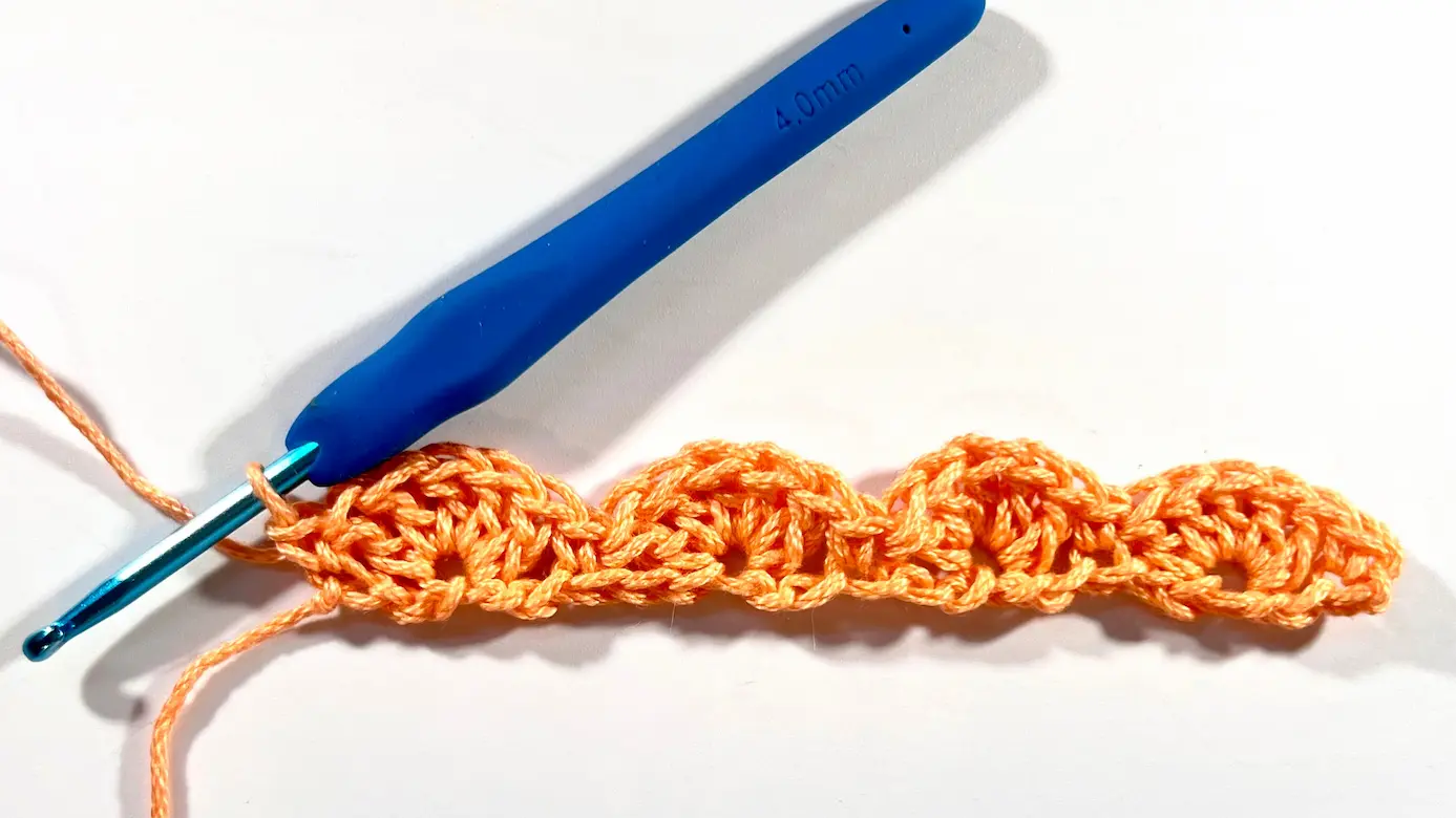 A completed first row of the shell stitch pattern in orange yarn, with a blue crochet hook inserted and ready to begin the next row.