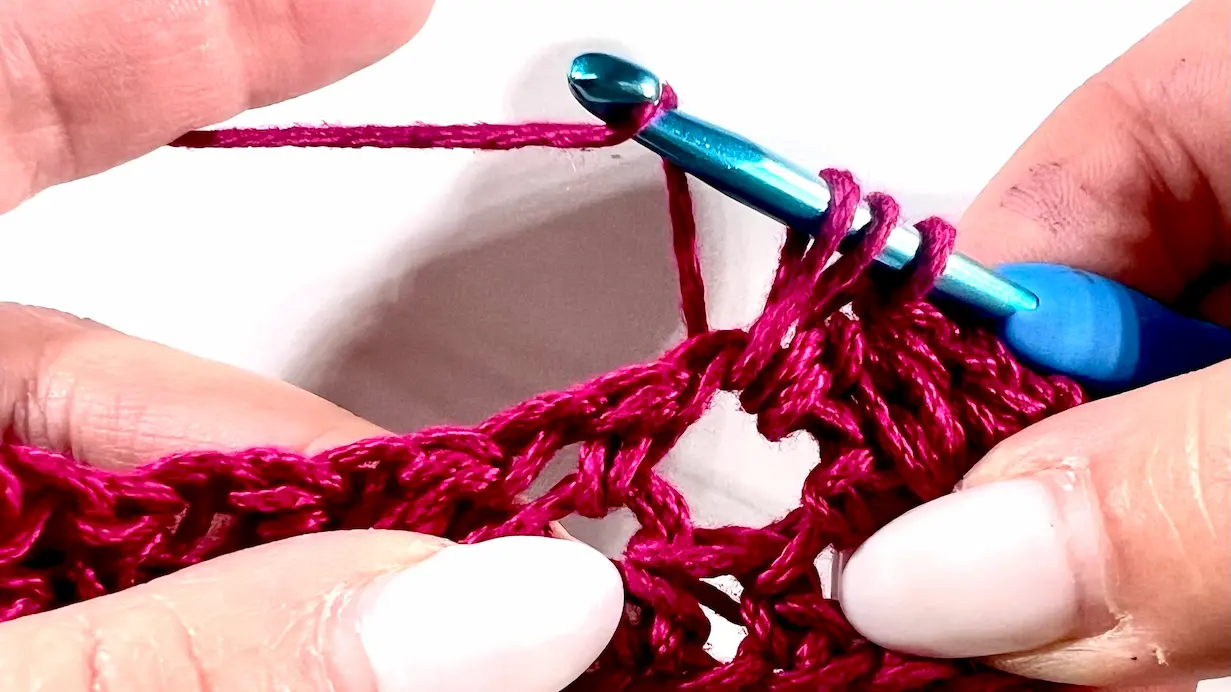 Close-up showing how to crochet the feather stitch step-by-step using fuchsia yarn and a blue crochet hook — part of a textured stitch tutorial.