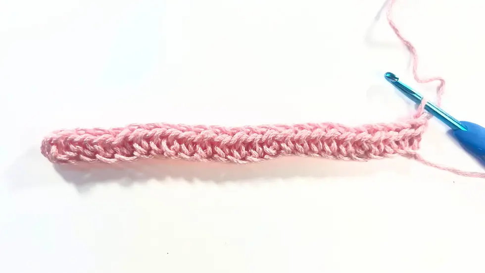 First row of half double crochet (HDC) stitches in pink yarn, forming the foundation for the camel stitch, with a crochet hook inserted into the last stitch on the right.