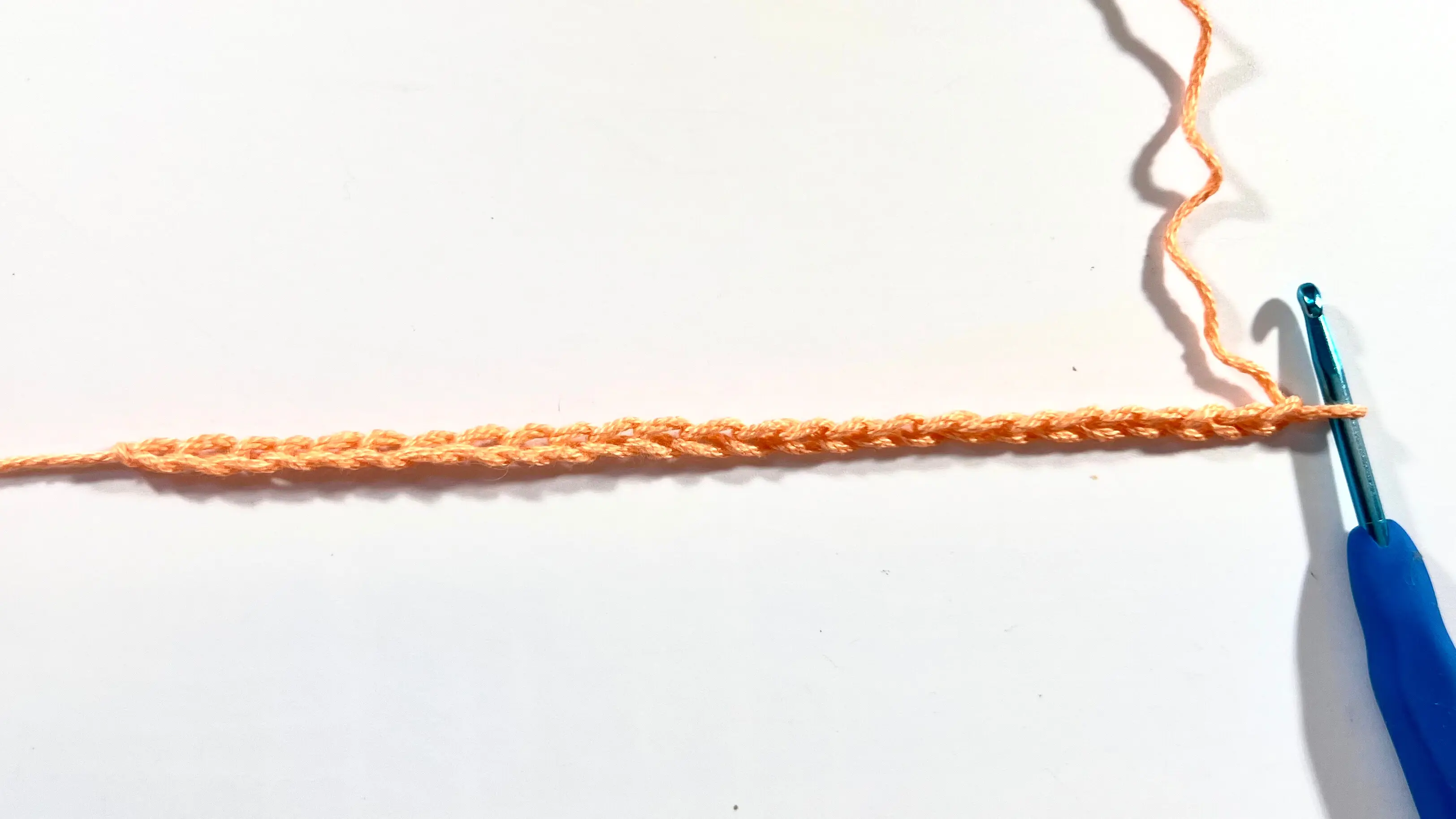 A close-up of an orange crochet foundation chain on a white background, with a blue crochet hook inserted into the last chain on the right.