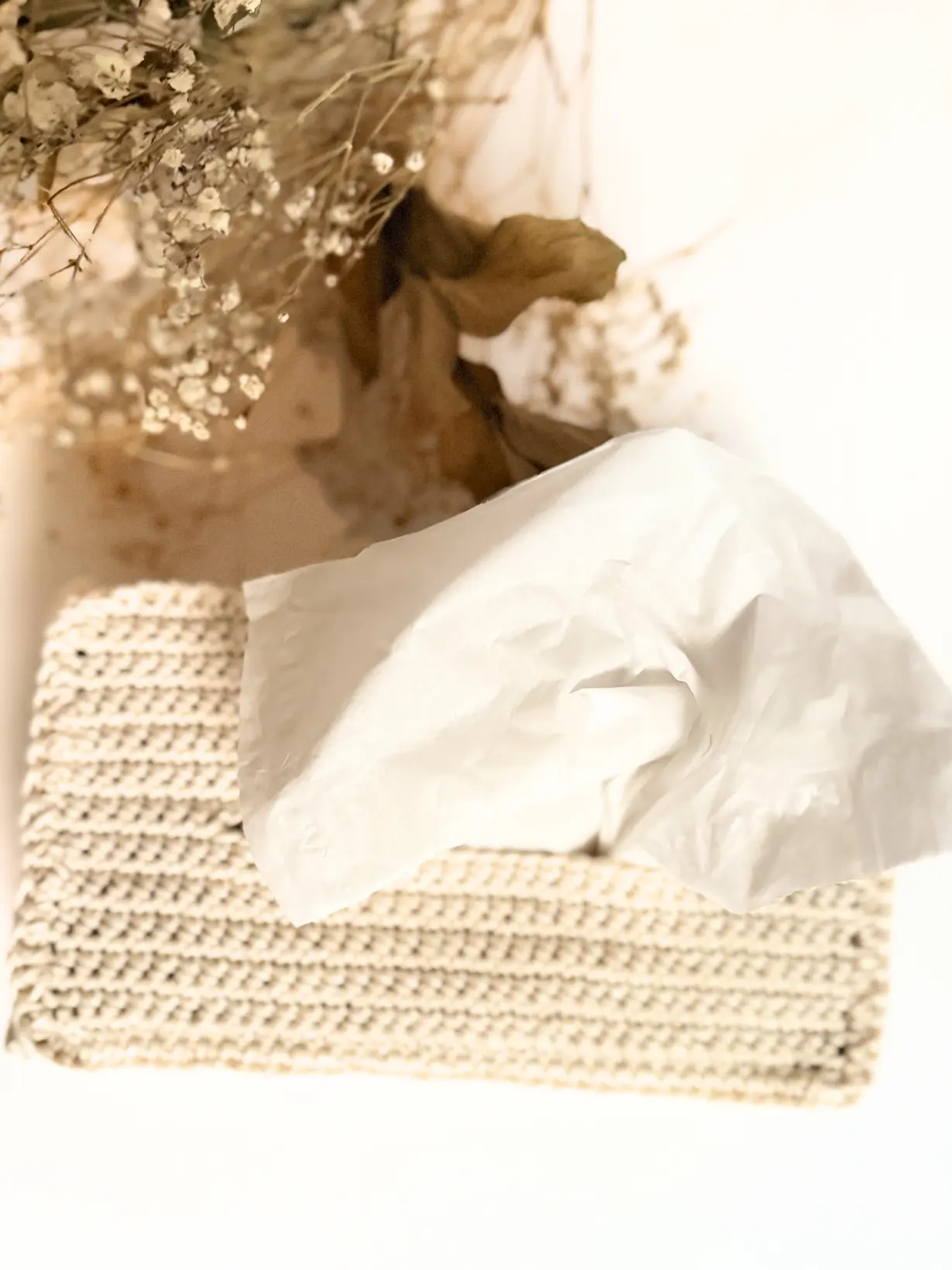 Close-up of a finished crochet tissue box cover with a tissue pulled through the opening and dried flowers in the background.