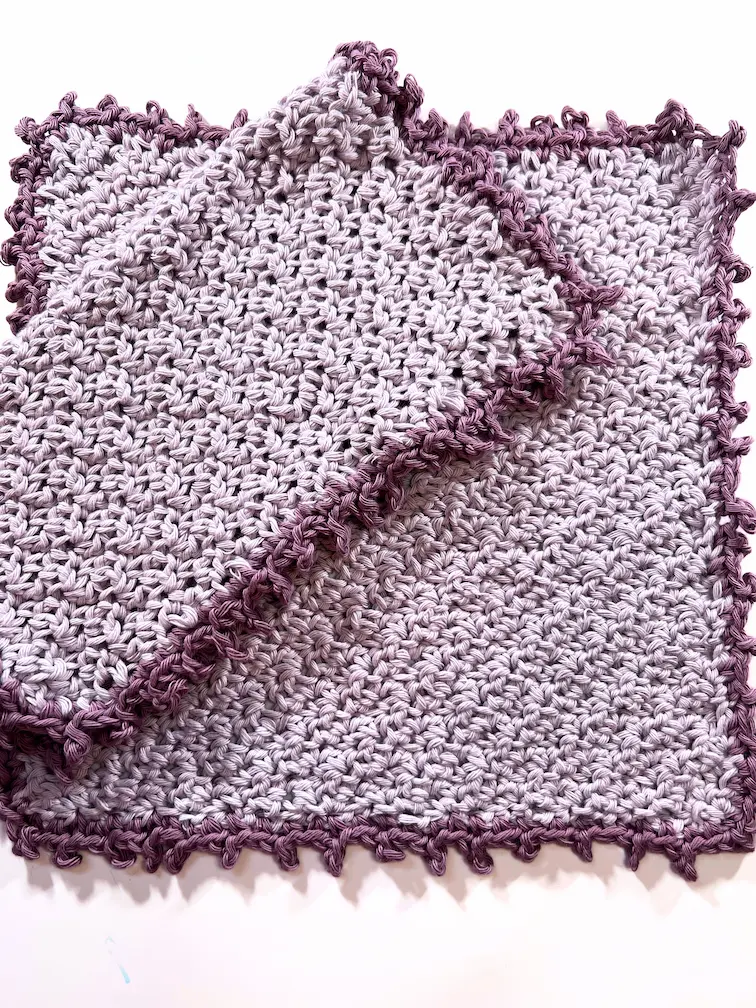 Two handmade crochet washcloths in light lavender yarn with a darker purple picot border, one folded over the other on a white background.