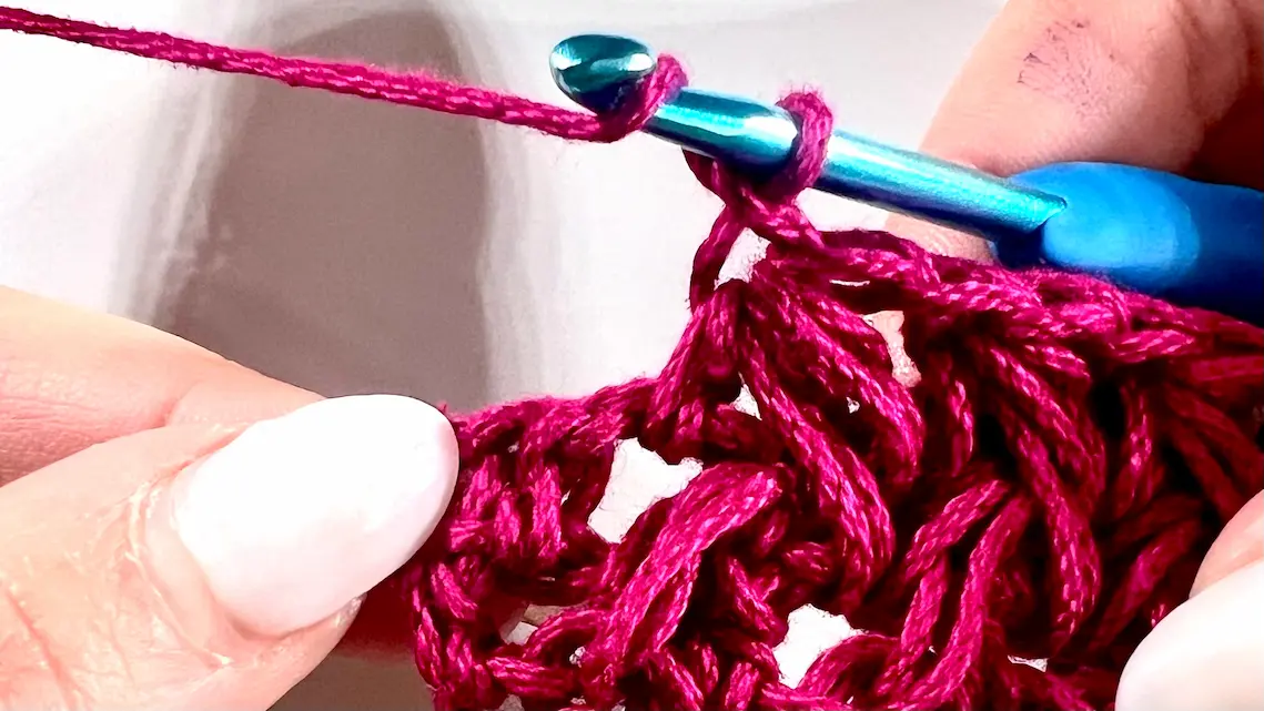 Close-up showing how to crochet the feather stitch step-by-step using fuchsia yarn and a blue crochet hook — part of a textured stitch tutorial.