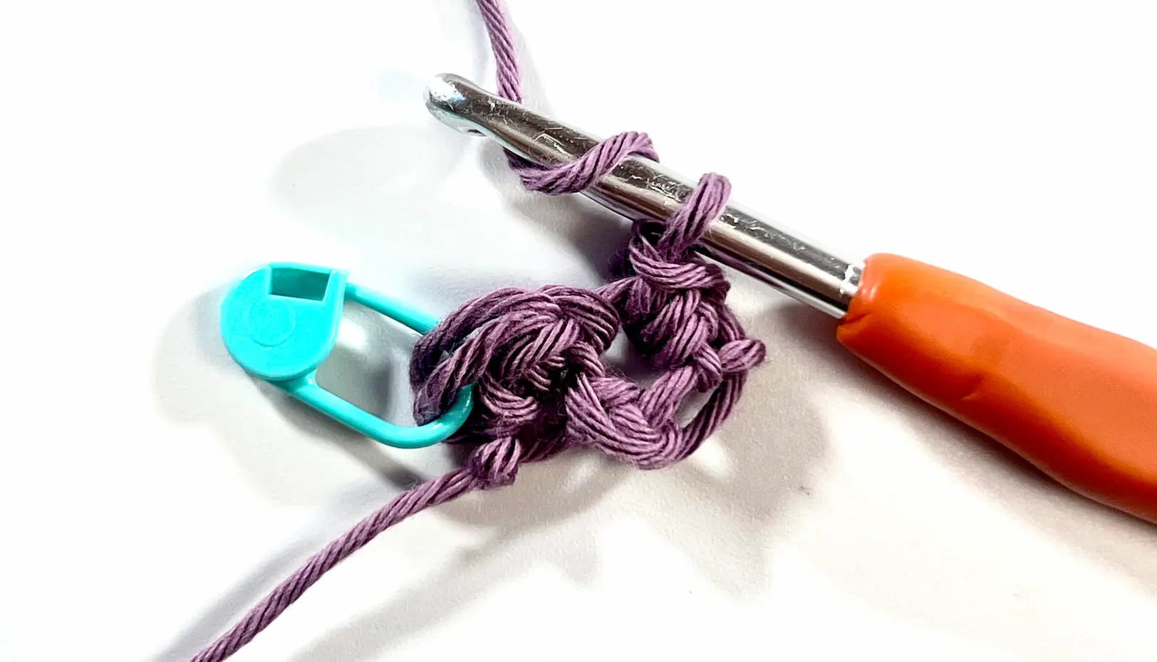 Crochet hook working the second block in C2C Even Moss Stitch. A teal stitch marker indicates the first stitch of the row, while a half double crochet is being formed on the next block with light purple yarn.