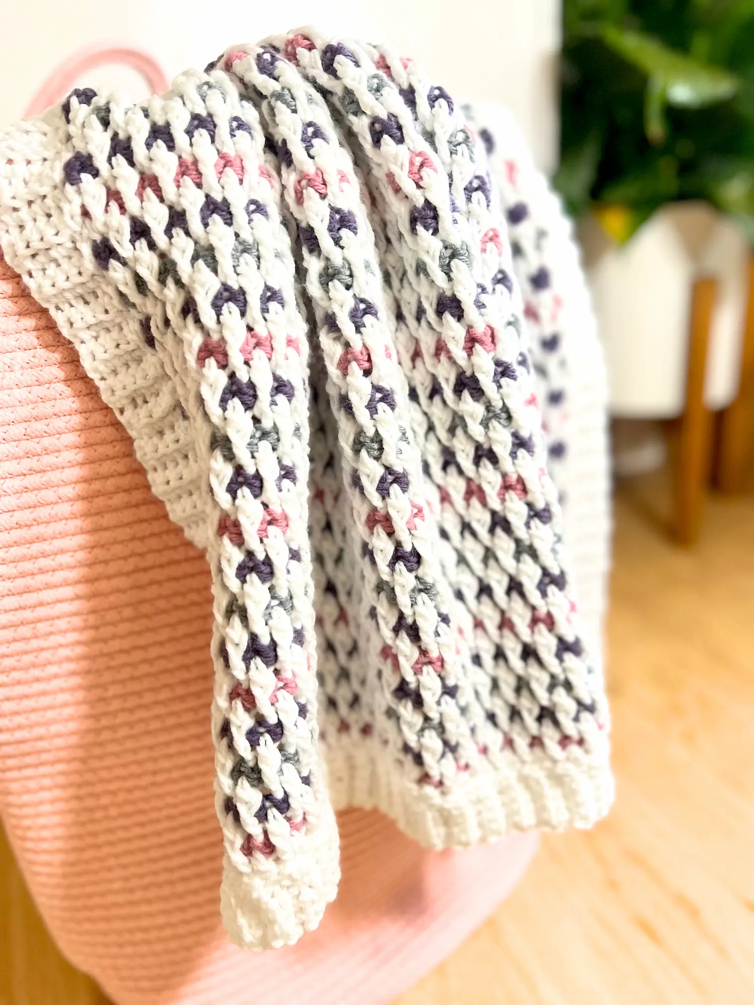 The Cuddle Blanket displayed over a pink basket, highlighting its soft texture, colour changes, and ribbed crochet border.