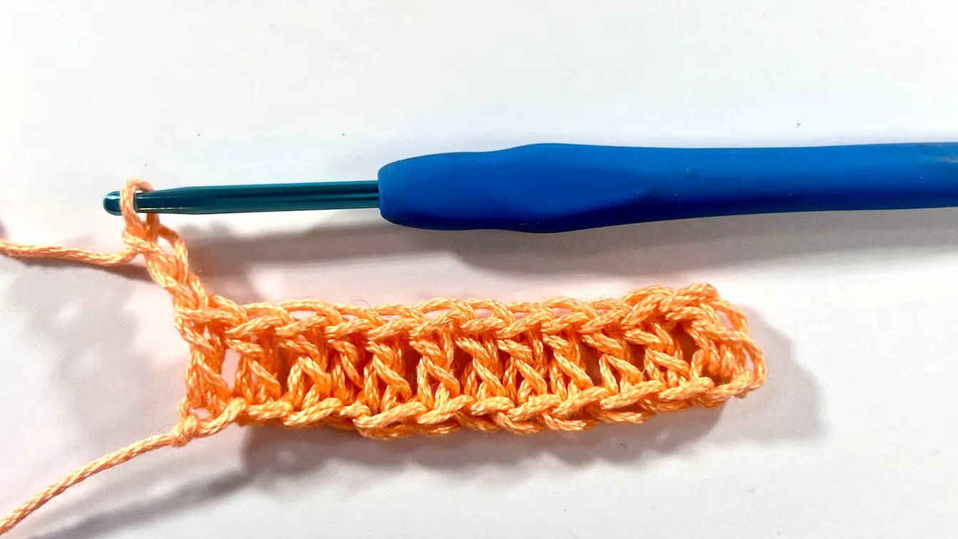 Row of double crochet stitches in orange yarn with a blue crochet hook, forming the waffle stitch base.