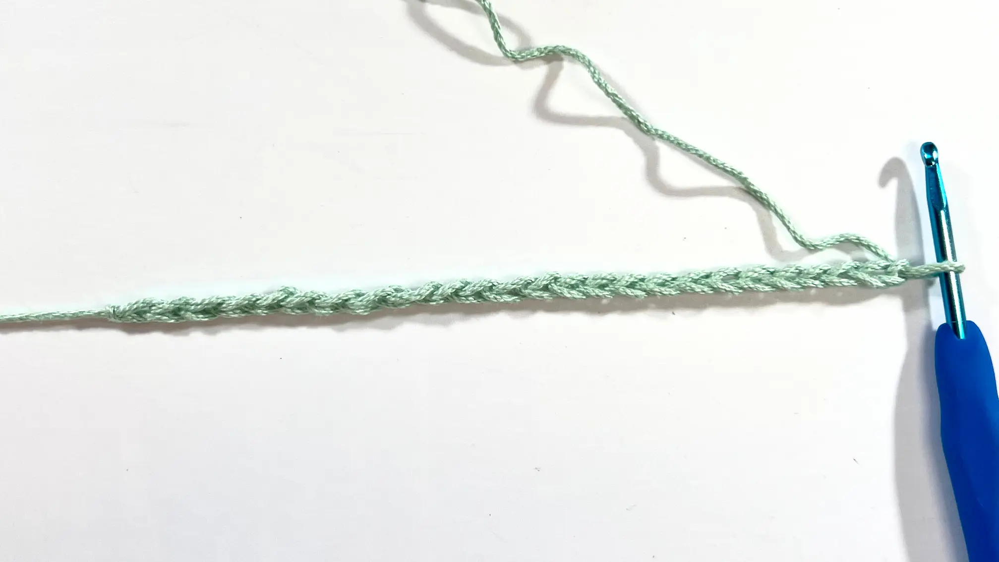 Foundation chain of light green yarn with crochet hook inserted, showing the starting point for the Crossed Double Crochet stitch.