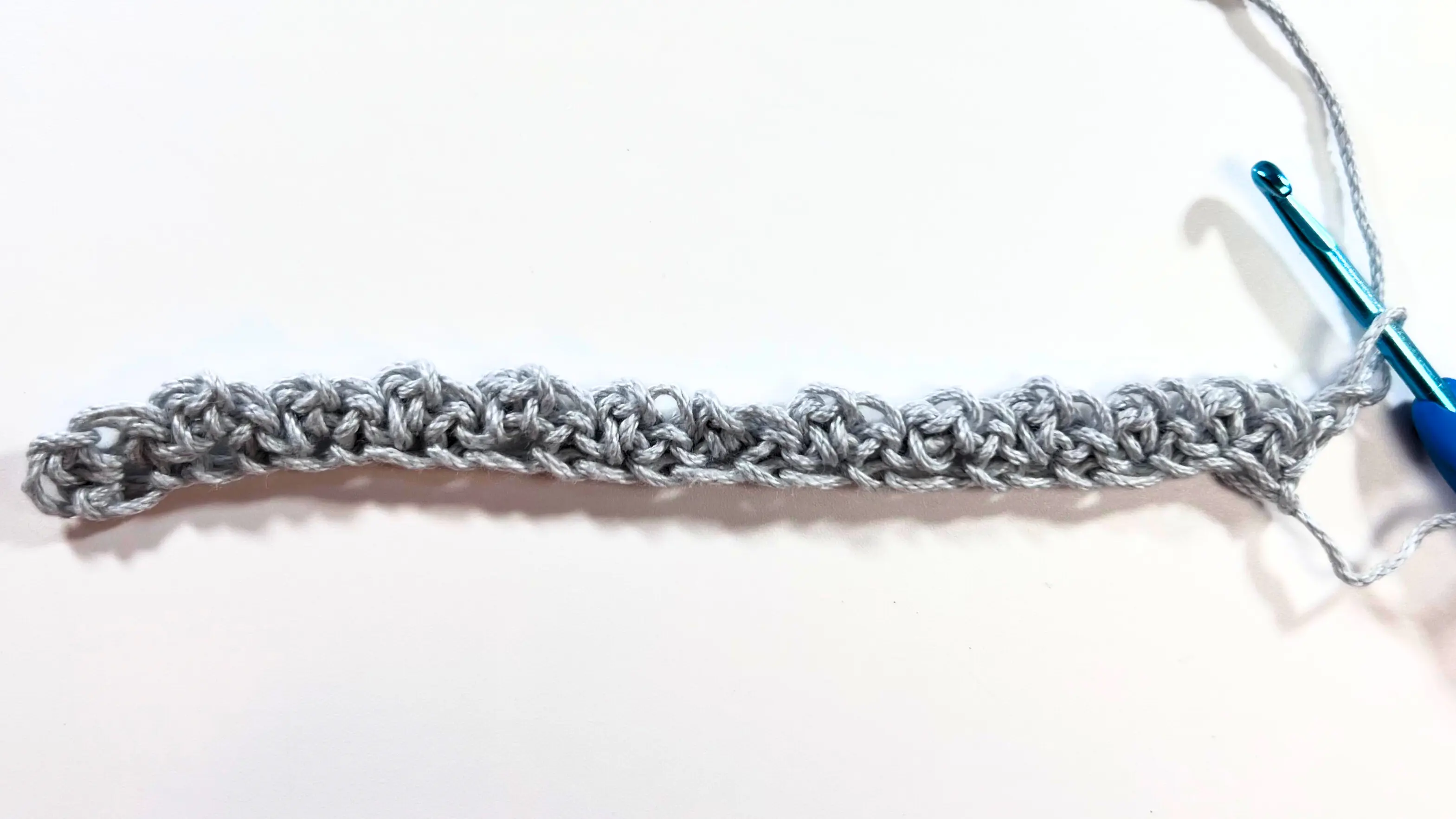 Full view of the finished first row of Spider Stitch, showing evenly spaced textured clusters across a light grey foundation chain, with the crochet hook in position to begin the next row.