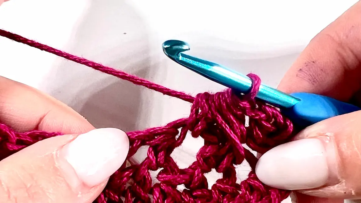 Close-up showing how to crochet the feather stitch step-by-step using fuchsia yarn and a blue crochet hook — part of a textured stitch tutorial.
