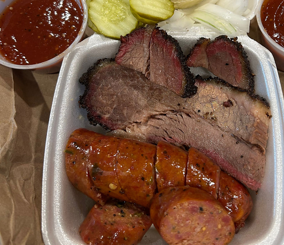 Sliced brisket and sausage in a tray with pickles, onions, and two cups of sauce, on a brown paper background.