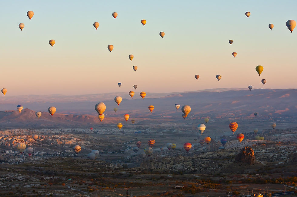 Many hot air balloons float over a rocky valley at sunrise, with pastel sky and distant mountains, creating a serene scene.