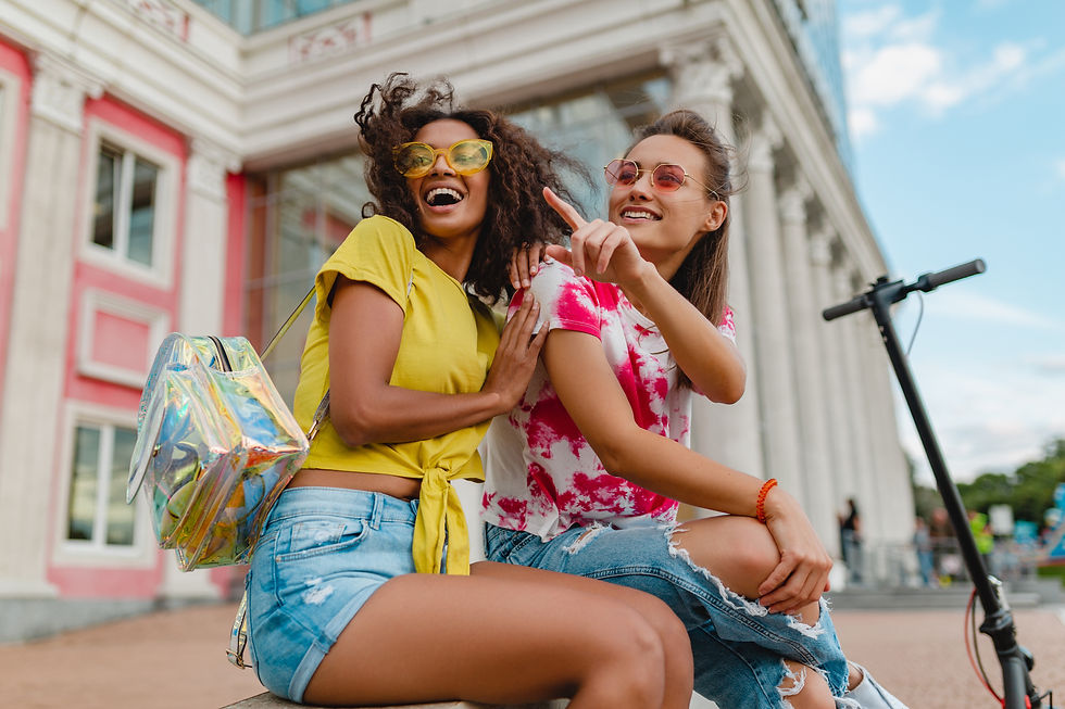 Two smiling women sit on a ledge outside a building. One points excitedly. They wear casual clothes and sunglasses. A scooter is nearby.