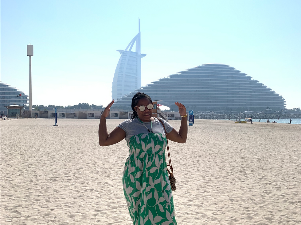 Woman in green dress poses on a sunny beach, with iconic Burj Al Arab hotel in the background. Clear blue sky, relaxed vibe.