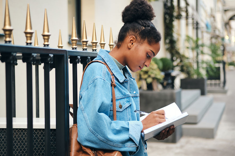 Young woman in a denim jacket intently writes in her notebook beside a decorative iron fence, capturing inspiration from the outdoor surroundings.