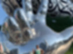 Large reflective sculpture of a hand with engraved patterns, set against a cityscape. A person poses below, flashing a peace sign.