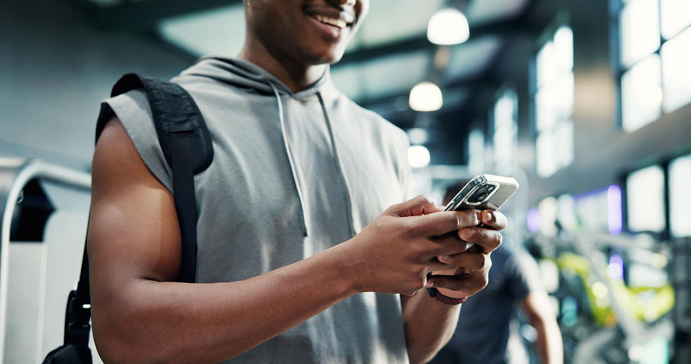 Man in a gray sleeveless hoodie smiles while using a smartphone in a bright gym. Background shows gym equipment and large windows.